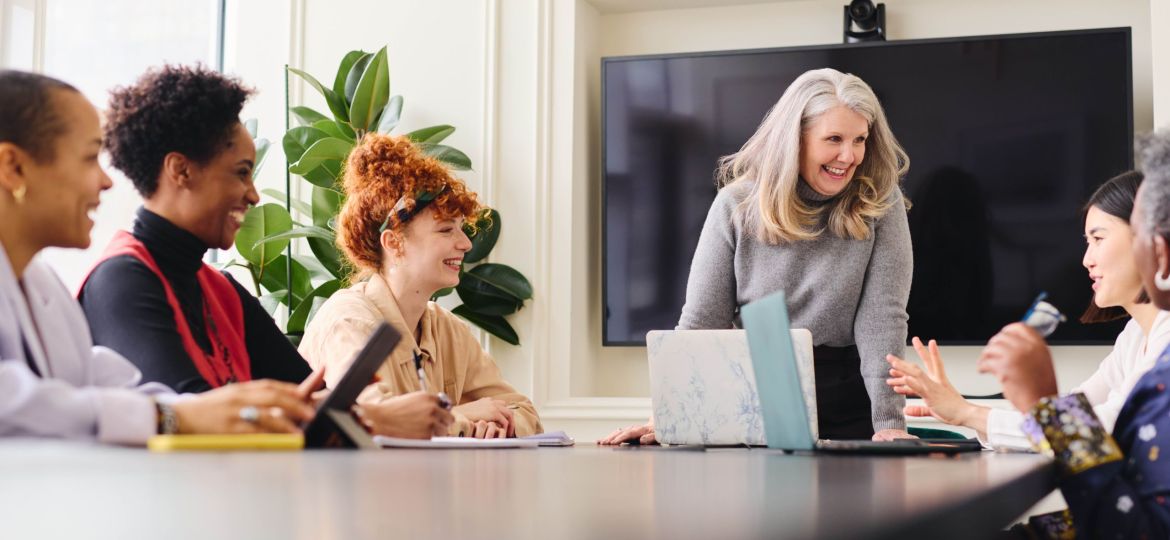 Portrait of senior white businesswoman with laptop smiling and listening to multiethnic female colleagues in boardroom